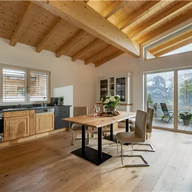 A modern dining room with wooden surfaces and large windows. In the background, you can see a beautiful mountain landscape.