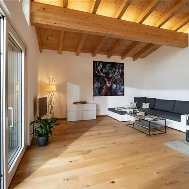 A modern living room with wooden ceilings and large windows. The furniture is simple and well arranged, featuring a comfortable sofa and a table.