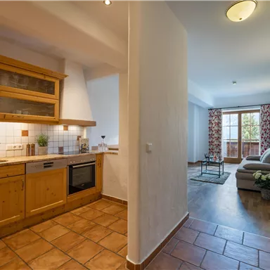 A modern kitchen with wooden shelves and a cozy seating area in the background. The room has tiled floors and access to a balcony with a window.