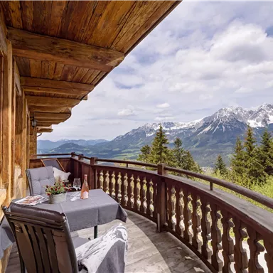 A beautiful balcony with a table and chairs, surrounded by an impressive mountain landscape. In the background, snow-covered peaks and green trees can be seen.