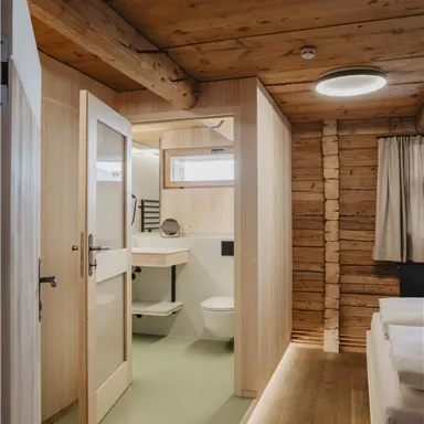 A modern, cozy room with wooden ceilings and warm light. The bathroom is bright and functional, separated by a door.
