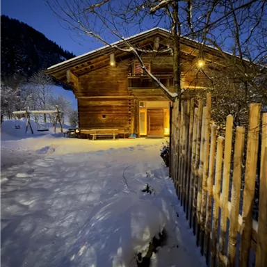 A cozy wooden house in the snow at night. Lights illuminate the surroundings and a fenced area is visible.
