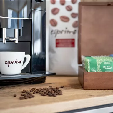 A coffee machine is on a wooden shelf with a cup. Next to it, there is a wooden box with various tea bags and coffee beans.