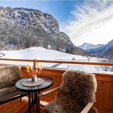 A cozy balcony with two chairs and a table, surrounded by snow-capped mountains. The sky is clear and the landscape is picturesque.