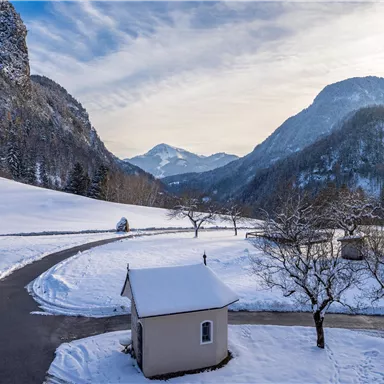 A snowy landscape with picturesque mountains in the background. A small, white building sits along a winding road, surrounded by trees.