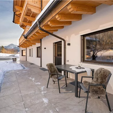 A modern terrace with two chairs and a table, surrounded by snow-capped mountains. The architecture combines wood and stone materials, creating a cozy atmosphere.