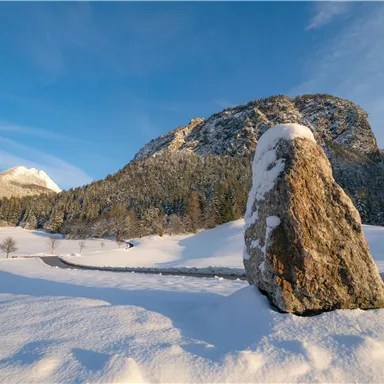 A winter landscape with snow-covered mountains and a large stone in the foreground. The sky is blue and clear.