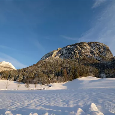 A winter landscape with snow-covered hills and a large rock in the background. The sky is clear and blue.
