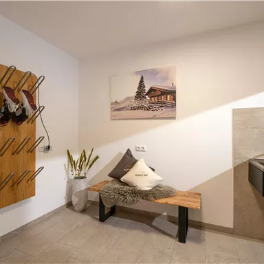 A modern entrance area with a shoe rack on the wall and a wooden bench. In the background, there is a sink and a picture of a chalet.