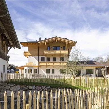 A modern alpine cabin with wooden cladding and a balcony. The garden is enclosed by a low wooden fence, surrounded by gentle hills.