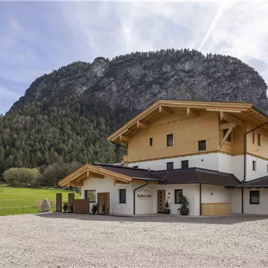 A modern building with wood and stone architecture is located in a picturesque setting. In the background, an impressive mountain rises with green forests.