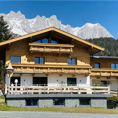 Ein gemütliches Holzhaus mit mehreren Balkonen und einer großen Terrasse. Im Hintergrund sind beeindruckende Berge und ein klarer blauer Himmel zu sehen.