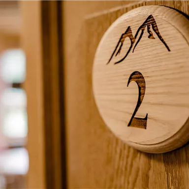 Ein hölzernes Türschild mit der Nummer 2 und einem Berg-Design. Im Hintergrund ist ein verschwommenes Zimmer zu sehen.