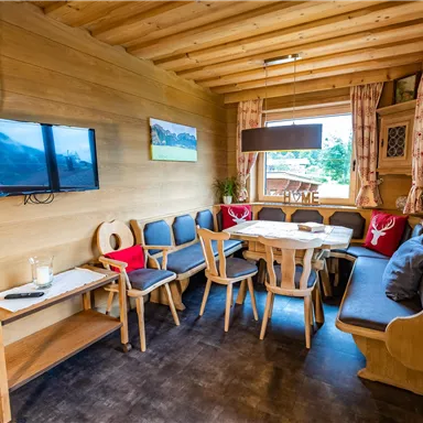 Ein gemütlicher Wohnraum mit Holzverkleidung und einer Sitzecke. Ein Fernseher hängt an der Wand, und ein großer Tisch lädt zum Essen ein.