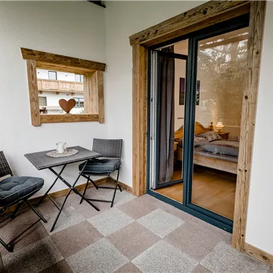 Ein gemütlicher Balkon mit zwei Stühlen und einem kleinen Tisch. Hinter einer Glas-Tür ist ein Schlafzimmer sichtbar.