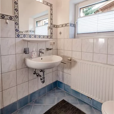Ein kleines Badezimmer mit weißen Fliesen und blauem Fußboden. Es gibt ein Waschbecken, Toiletten und ein Fenster mit Licht.
