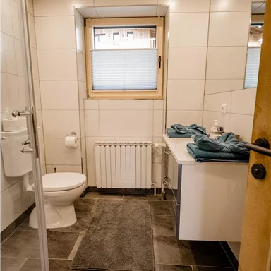 Ein modernes Badezimmer mit einer Dusche, einem WC und einem Waschbecken. Handtücher in Türkisfarbe liegen bereit, und ein Fenster sorgt für Tageslicht.