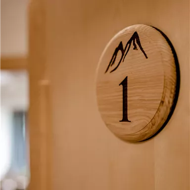 Ein hölzerner Türschild mit der Nummer 1 und einem Bergsymbol. Der Hintergrund ist unscharf und zeigt einen Bereich drinnen.