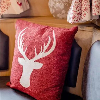 Ein rotes Kissen mit dem Motiv eines Rehkodies liegt auf einer dunklen Couch. Im Hintergrund sind gemusterte Vorhänge und eine dekorative Lampe sichtbar.