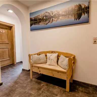 Ein einladender Flur mit einem Holzsofa und Kissen. An der Wand hängt ein großes Bild von einer Berglandschaft mit gespiegelt ruhig im Wasser.