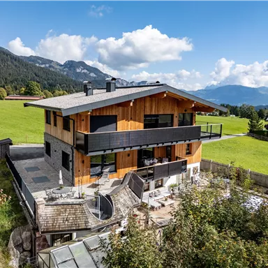 Ein modernes Holzhaus in einer malerischen Landschaft. Im Hintergrund sind Berge und ein blauer Himmel mit Wolken zu sehen.