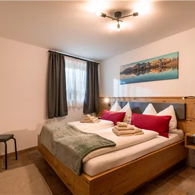 Ein modernes Schlafzimmer mit einem Doppelbett und holzfarbenen Möbeln. Der Raum ist hell mit einem großen Fenster und einem Fernseher an der Wand.
