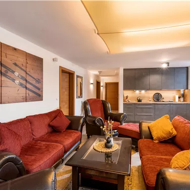 Ein modernes Wohnzimmer mit roten Sofas und einem eleganten Tisch. Die offene Küche ist im Hintergrund sichtbar und sorgt für eine gemütliche Atmosphäre.