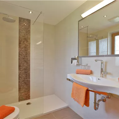 Ein modernes Badezimmer mit einer Dusche und einem Waschbecken. Orangefarbene Handtücher und eine helle, freundliche Atmosphäre schaffen ein einladendes Ambiente.