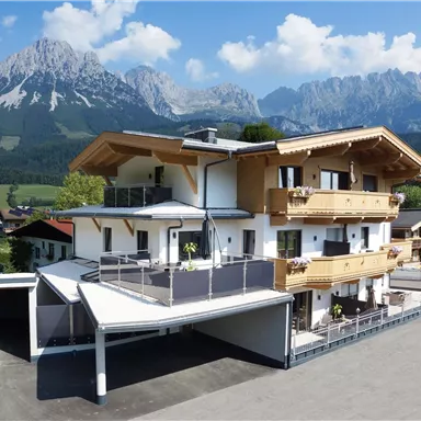 A modern chalet in Alpine style with large balconies. In the background, majestic mountains and a green landscape can be seen.
