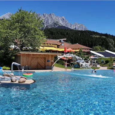 Ein schöner Pool mit klarem Wasser und einer sonnigen Umgebung. Im Hintergrund sind Berge und Grünflächen zu sehen.