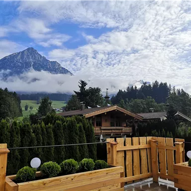 Eine malerische Berglandschaft mit hohen Gipfeln und bewölktem Himmel. Im Vordergrund eine Holzterrasse mit grünen Pflanzen und Blick auf angrenzende Häuser.