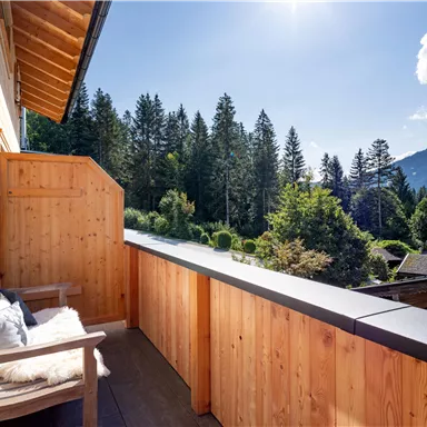 Ein Holzbalkon mit einem gemütlichen Sitzplatz. Im Hintergrund sind grüne Wälder und Berge zu sehen.