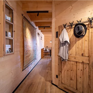Ein gemütlicher Flur mit Holzvertäfelungen und einer hölzernen Tür. An der Wand hängen ein Mantel und ein Hut, während ein weiteres Zimmer im Hintergrund sichtbar ist.