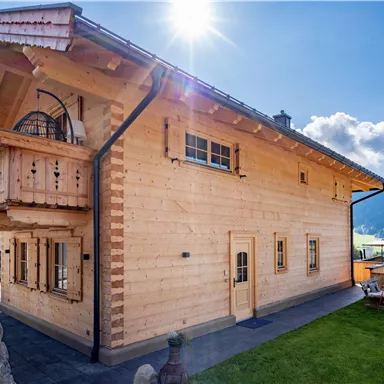 Ein modernes Holzhaus mit einem Balkon und großen Fenstern. Die Umgebung ist grün und hügelig, mit einem strahlend blauen Himmel.