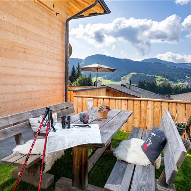 Eine gemütliche Terrasse mit einem Holztisch und Bänken, umgeben von einer schönen Berglandschaft. Im Hintergrund sind grüne Wiesen und ein blauer Himmel mit Wolken zu sehen.