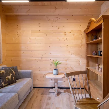 Ein gemütlicher Raum mit einer grauen Couch, einem kleinen Tisch und einem Holzregal. Holzpaneele an den Wänden schaffen eine warme Atmosphäre.