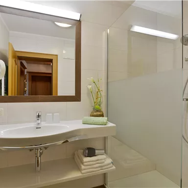 Ein modernes Badezimmer mit Dusche und großem Spiegel. Auf dem Waschbecken liegen frische Handtücher und Pflanzen als Dekoration.