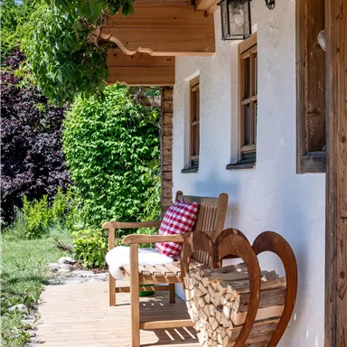 Eine gemütliche Veranda mit einem Holzstuhl und einem Herzen aus Holz für Holzscheite. Im Hintergrund sind grüne Pflanzen und ein schöner Garten zu sehen.