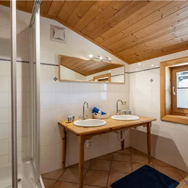 Ein modernes Badezimmer mit einer Dusche und einem Doppelwaschbecken. Holzdecken und große Fenster sorgen für eine helle, freundliche Atmosphäre.