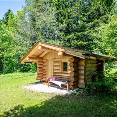 Eine gemütliche Blockhütte steht in einer grünen Landschaft. Umgeben von Bäumen bietet sie einen ruhigen Rückzugsort.