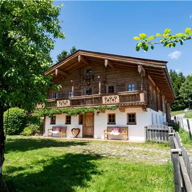 Ein traditionelles Holzhaus mit Balkon in einer grünen Umgebung. Es ist ein sonniger Tag mit blauem Himmel und Bäumen im Vordergrund.