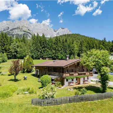 Eine malerische alpine Landschaft mit einem Holzhaus und üppigem Grün. Im Hintergrund erheben sich beeindruckende Berge unter einem blauen Himmel.