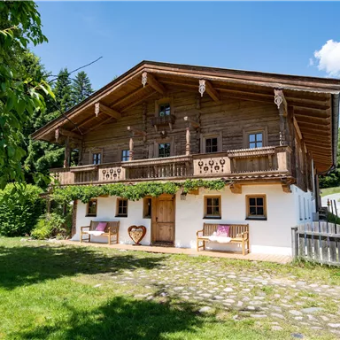 Ein traditionelles Holzhaus im alpenländischen Stil mit einem großen Balkon. Umgeben von saftigem Grün und Bäumen, vermittelt es eine gemütliche Atmosphäre.