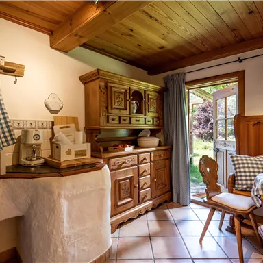 Eine gemütliche Küche mit Holzschränken und einem großen Fenster, das Licht hereinlässt. Der Raum ist mit einem karierten Tischdecken und landhausstilistischen Dekorationen ausgestattet.