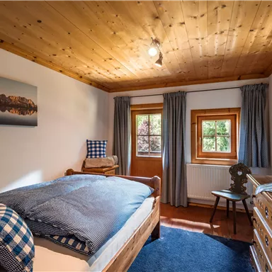 Ein gemütliches Schlafzimmer mit Holzdecke und großen Fenstern. Es gibt ein Einzelbett, einen Stuhl und eine Kommode sowie eine Landschaftsaufnahme an der Wand.