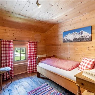 Ein gemütliches Zimmer mit Holzwänden und einem Einzelbett. Rote kariert Vorhänge und ein schöner Ausblick auf die Berge ergänzen die rustikale Atmosphäre.