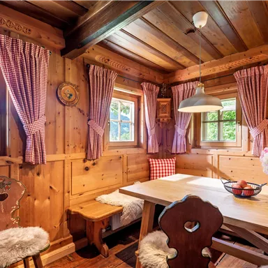 Ein gemütliches Holzhäuschen mit einem rustikalen Esstisch und Stühlen. Die Wände sind aus Holz, und die Fenster sind mit karierten Vorhängen geschmückt.