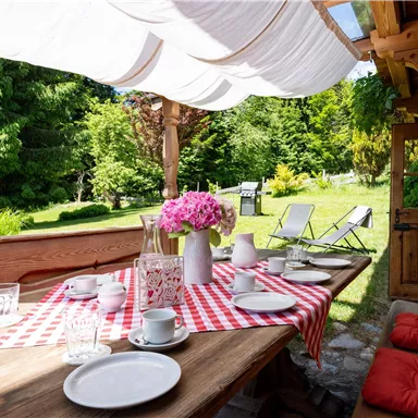 Ein einladender Esstisch im Freien mit rotem Karomuster. Der Tisch ist mit Geschirr und frischen Blumen dekoriert, umgeben von einem grünen Garten.