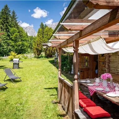 Eine charmante Terrasse mit Holzmöbeln und blühenden Blumen. Im Hintergrund sieht man eine grüne Wiese mit Liegen und üppiger Vegetation.