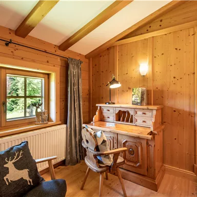 Ein gemütliches Holzinterieur mit einem Schreibtisch, einem Stuhl und einem Fenster, das auf einen Gartenblick zeigt. Eine Wanduhr ergänzt die warme Atmosphäre des Raums.